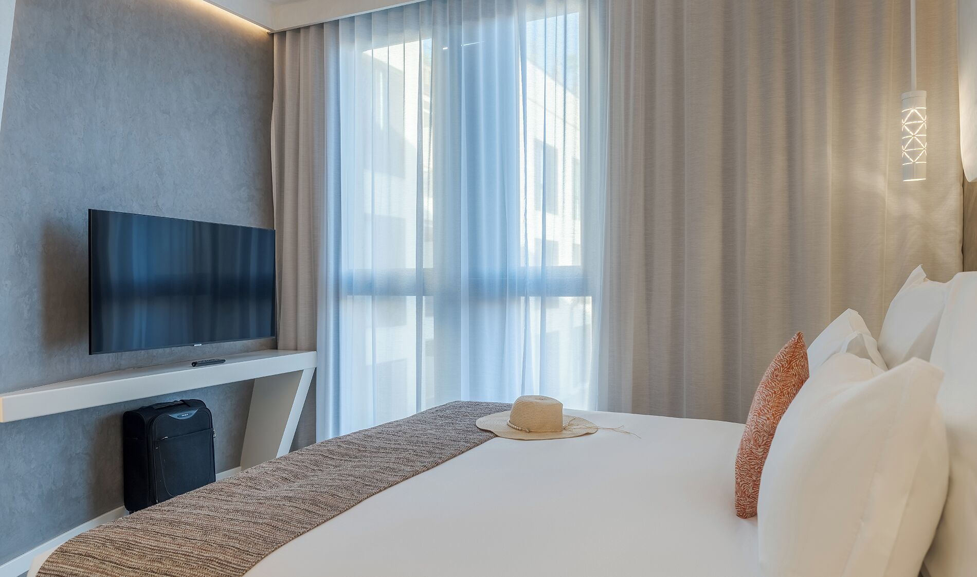 A hotel room with a large bed, a flat-screen TV mounted on the wall, and a window with sheer curtains.