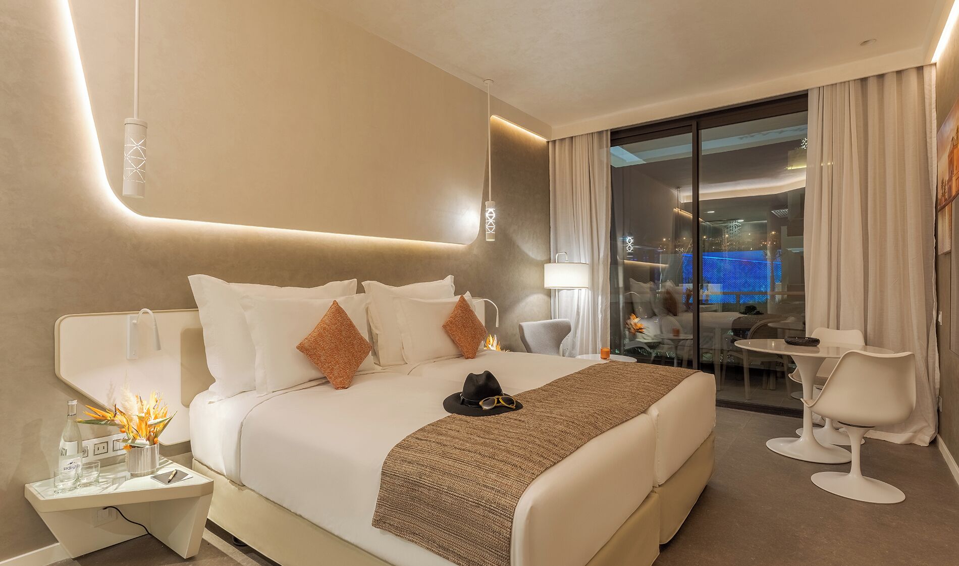 A modern hotel room with a large bed, a sitting area with a table and chairs, and a view of a cityscape through the glass door.