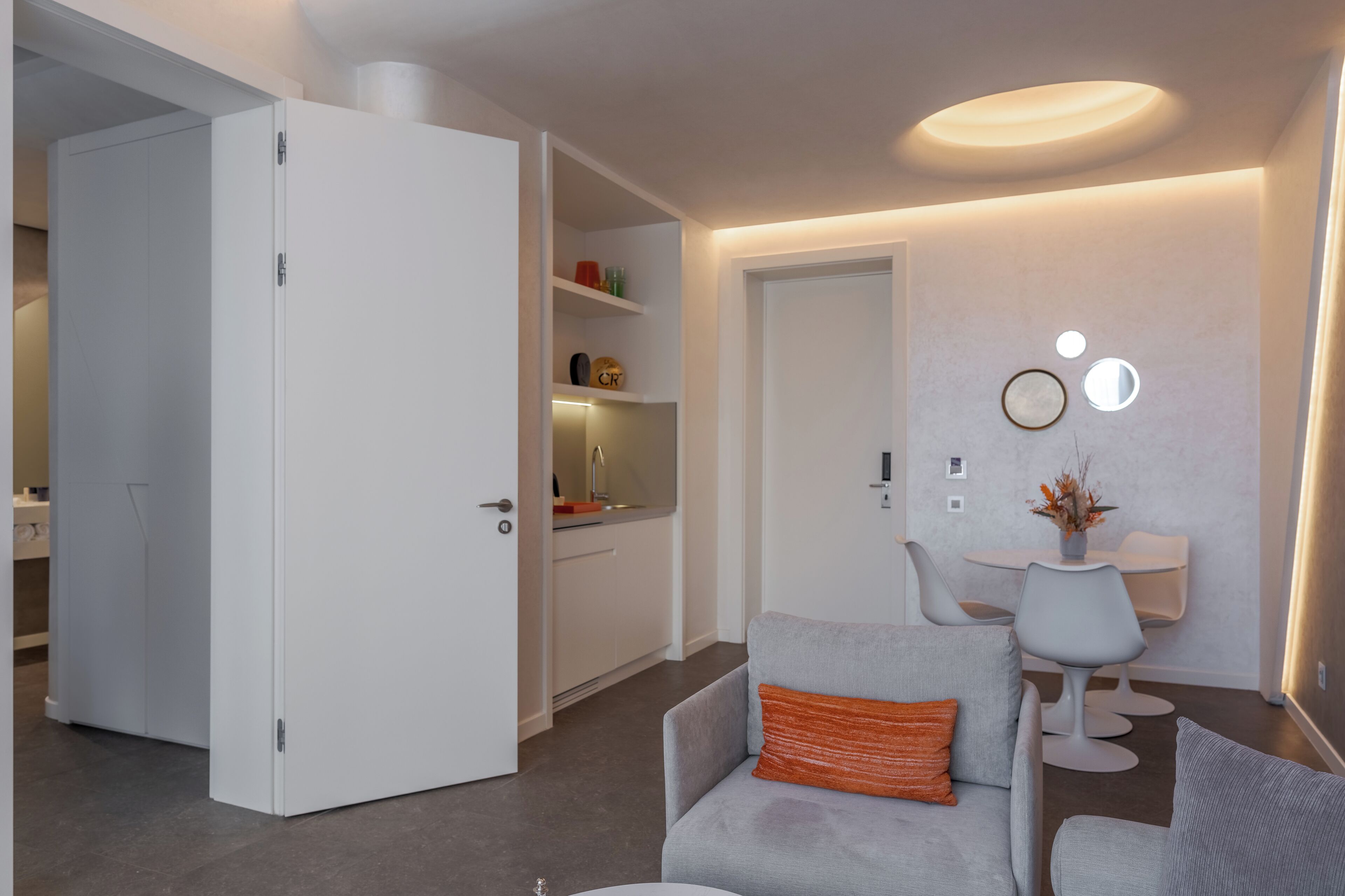 A modern living room with a grey sofa, a small table with chairs, and a kitchen area visible through an open door.