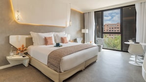 Una habitación de hotel moderna con una cama grande, mesitas de noche, un escritorio y un balcón con vistas a edificios.
