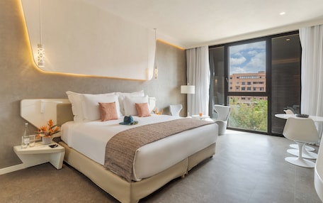 A modern hotel room with a large bed, bedside tables, a desk, and a balcony with a view of buildings.