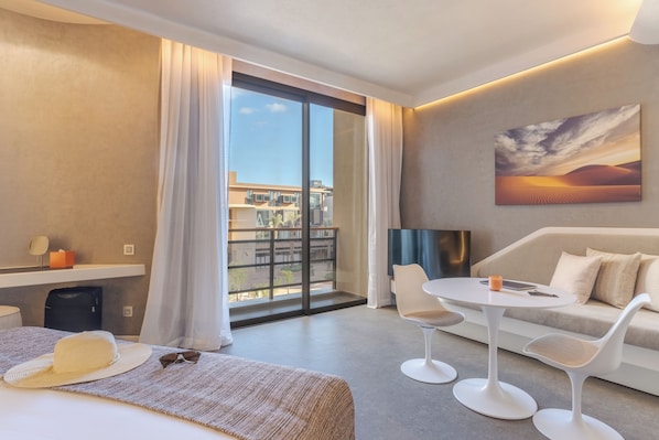 Una habitación de hotel moderna con un amplio ventanal corredero al balcón, un sofá, dos sillas blancas, una mesita y un cuadro en la pared.