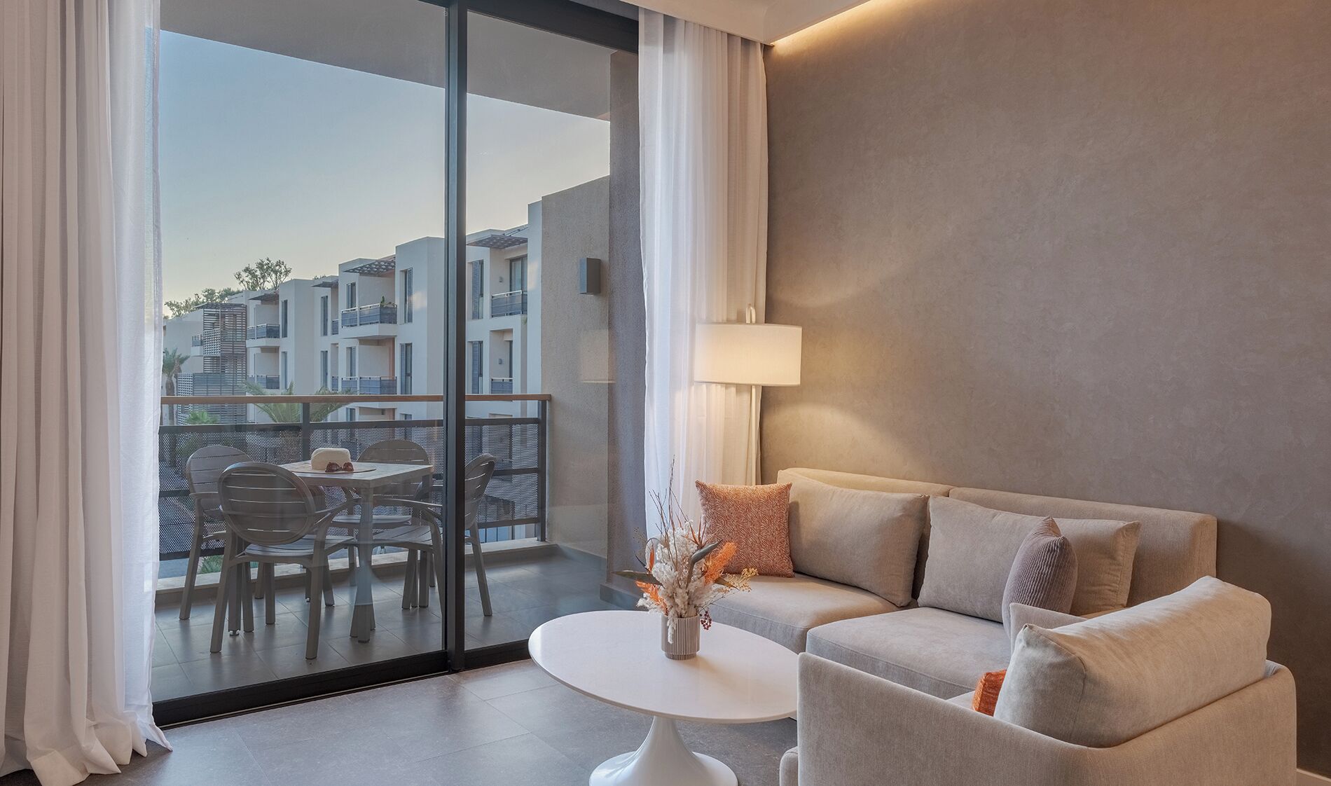Una habitación de hotel moderna con un balcón, un sofá, una pequeña mesa redonda y una lámpara.