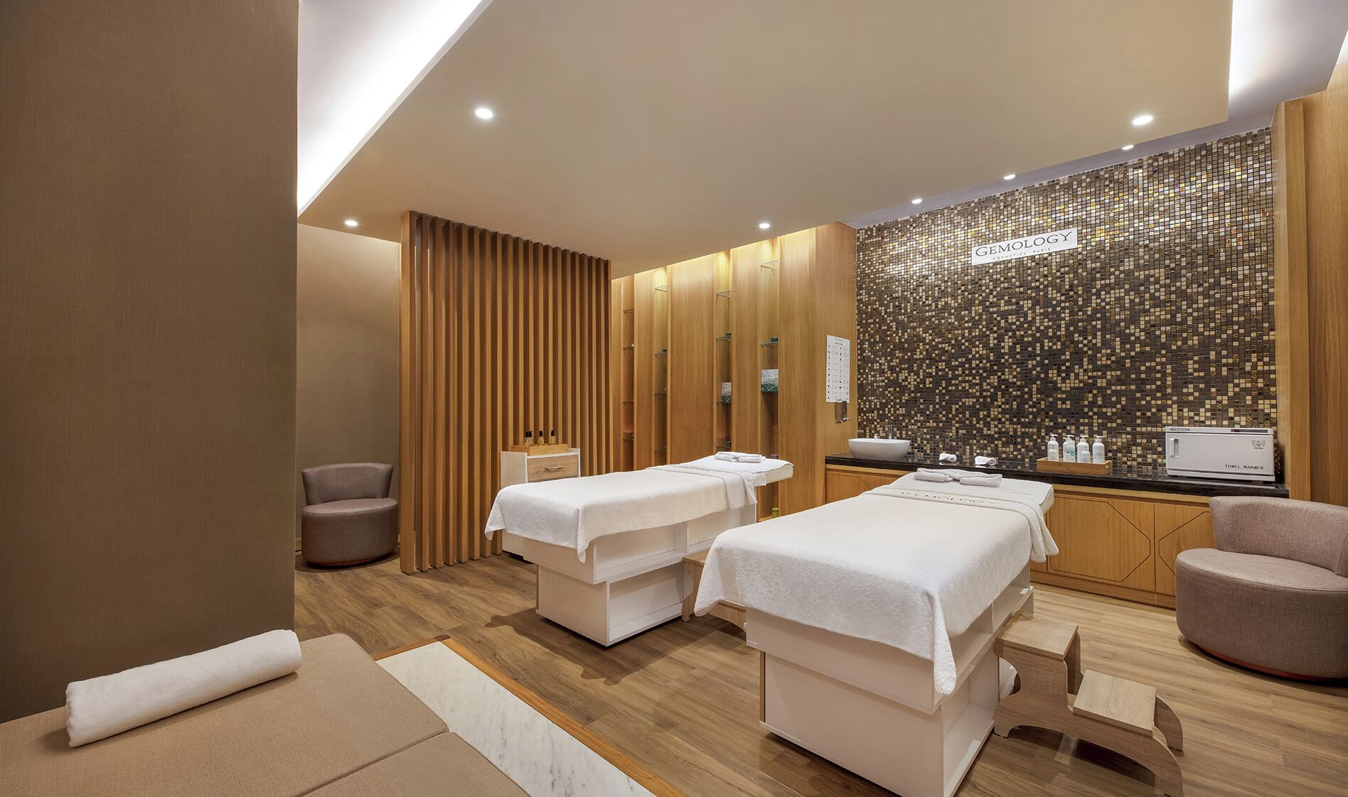 A modern spa room with two massage tables, wooden paneling, a tiled mosaic wall, and a reception area with a computer and toiletries.