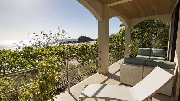 Een balkon met een witte lounge stoel, beklede zitplaatsen en uitzicht op de oceaan en een rotsformatie.