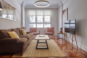 Een woonkamer met een bruine bank, twee fauteuils, een salontafel, een tv op een dressoir, en grote ramen.