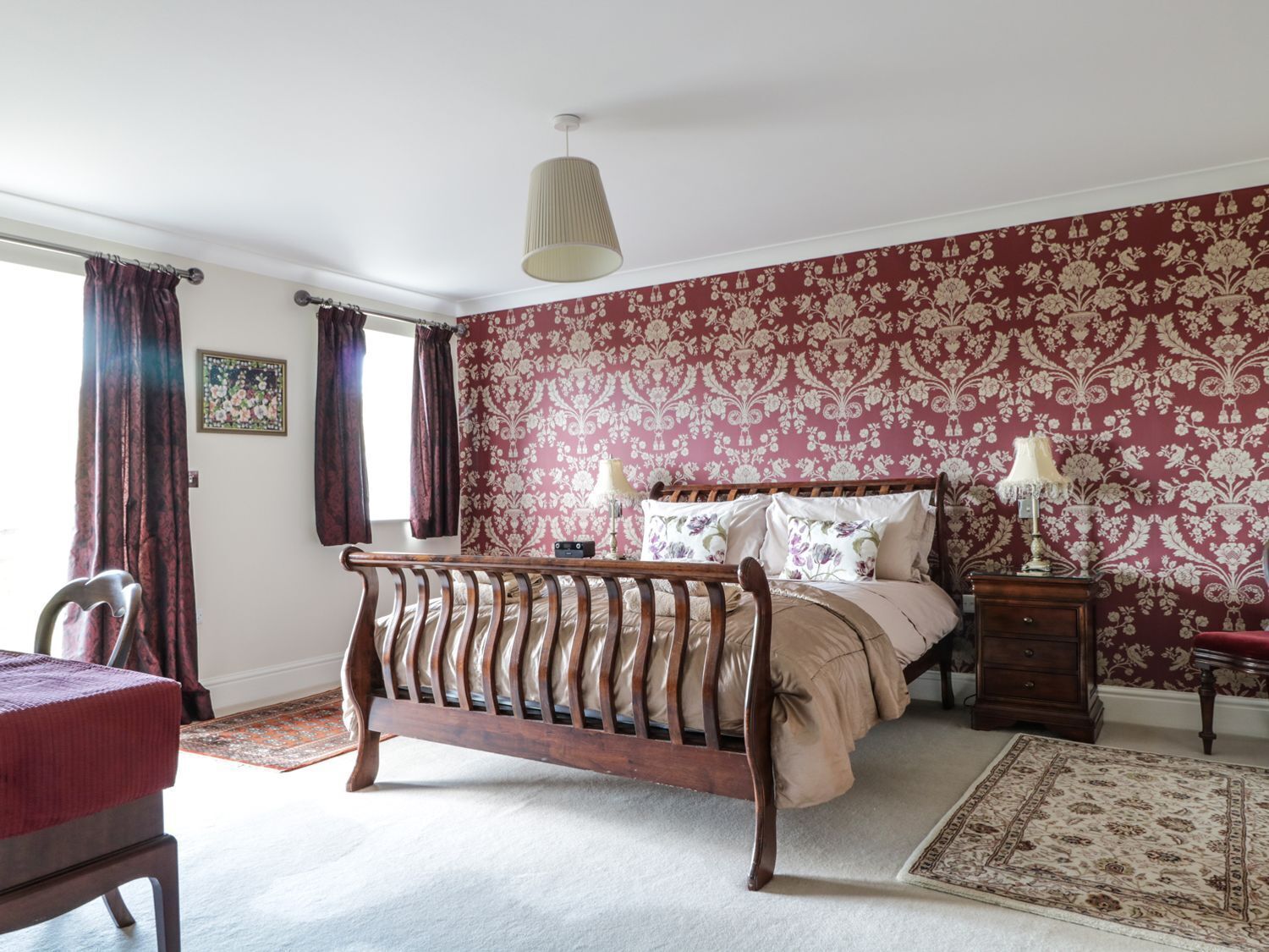 A bedroom with a wooden bed, floral wallpaper, a red chair, and a small bedside table with a lamp.