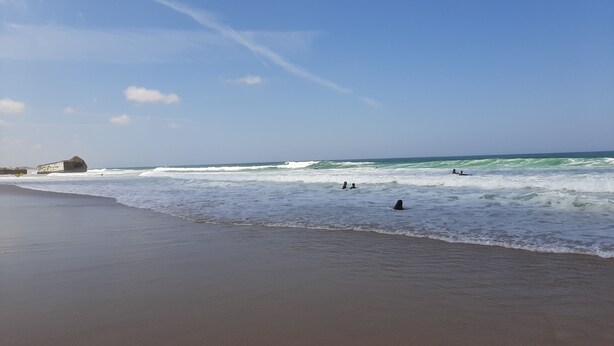 Ein Strand mit Wellen, blauem Himmel und einem großen Felsen links.