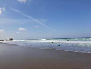 A beach with waves, a clear sky, and a large rock on the left.