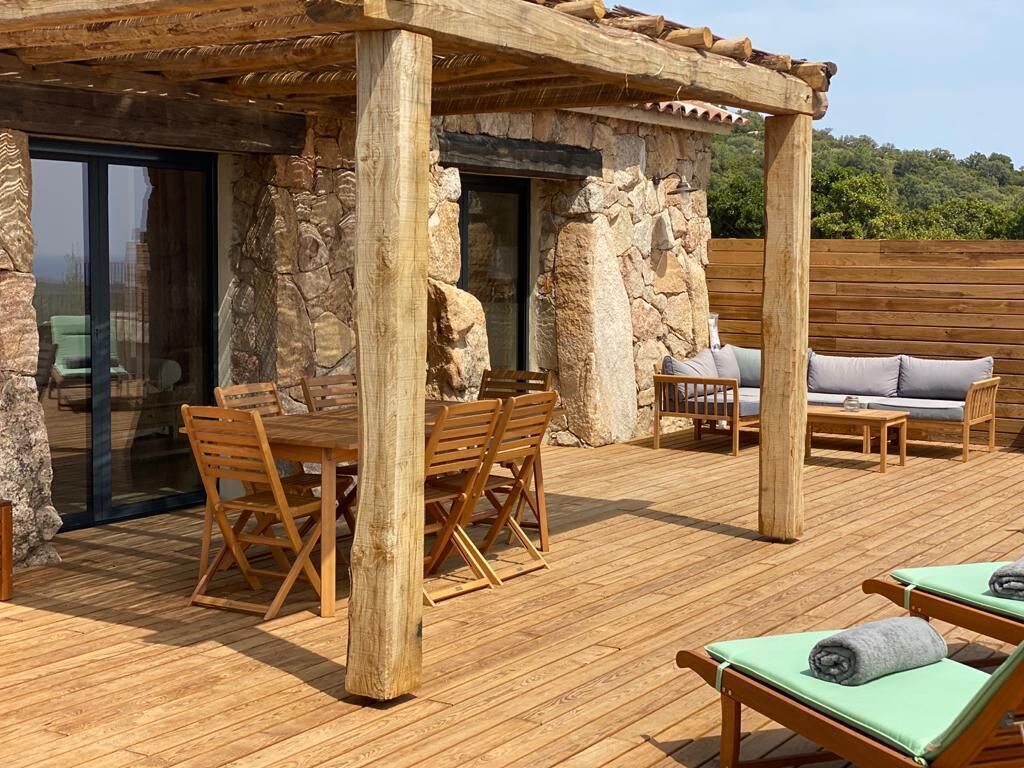 An outdoor wooden deck with a stone wall, a table with chairs, a sofa, and lounge chairs.