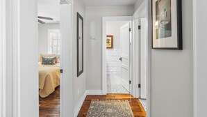 Un couloir avec un plancher en bois, des murs blancs et un tapis. On y trouve une porte qui mène à une salle de bain dont un mur est recouvert de carreaux blancs et sur lequel est accroché un tableau encadré.