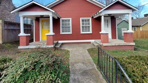 Une maison à deux étages, avec une façade rouge, des éléments décoratifs blancs et une porte d’entrée jaune. Elle possède deux petits porches dotés de colonnes blanches et de piliers en briques.