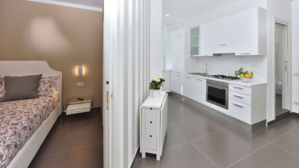 Een moderne slaapkamer met een bed, nachtkastje en een klein tafeltje. Grenzend aan de slaapkamer bevindt zich een keuken met witte kasten, een fornuis en een spoelbak.