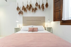 Un lit bien fait, recouvert d’une couvre-lit rose à motifs, avec une tête de lit beige et une composition florale suspendue au-dessus. On y trouve deux tables de chevet avec des lampes, ainsi qu’une petite table de nuit avec une bouteille.