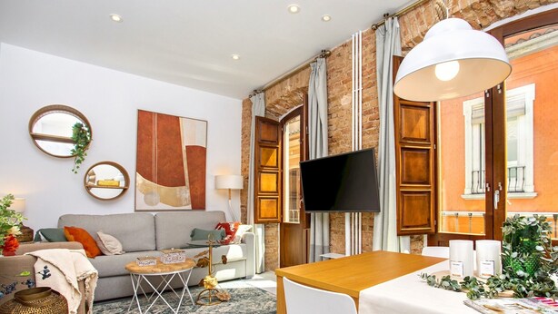 Ein modernes Wohnzimmer mit Sofa, Couchtisch und Fernseher. Es gibt einen Essbereich mit Tisch und Stühlen. Der Raum hat eine Backsteinwand und große Fenster.