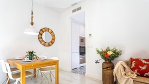 Ein modernes Wohnzimmer mit einem Holzesstisch, weißen Stühlen, einer hängenden Pendelleuchte, einem runden Spiegel und einem Sofa mit einem roten Kissen.