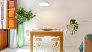 A dining area with a wooden table, two chairs, a hanging light fixture, a potted plant, and a wall-mounted shelf with greenery.
