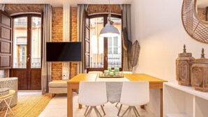 A dining area with a wooden table, two white chairs, a TV mounted on the wall, and a view of a balcony through large windows.