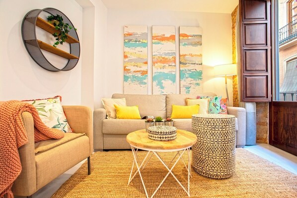 A living room with a sofa, a round coffee table, and a patterned ottoman. There are three abstract paintings on the wall and a lamp on a side table.