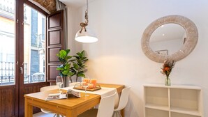 Un comedor con una mesa de madera, sillas blancas, una lámpara colgante, un espejo redondo y un estante blanco.