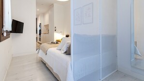 Un dormitorio con cama, televisión montada en la pared, mesita de noche con lámpara y un espejo grande.