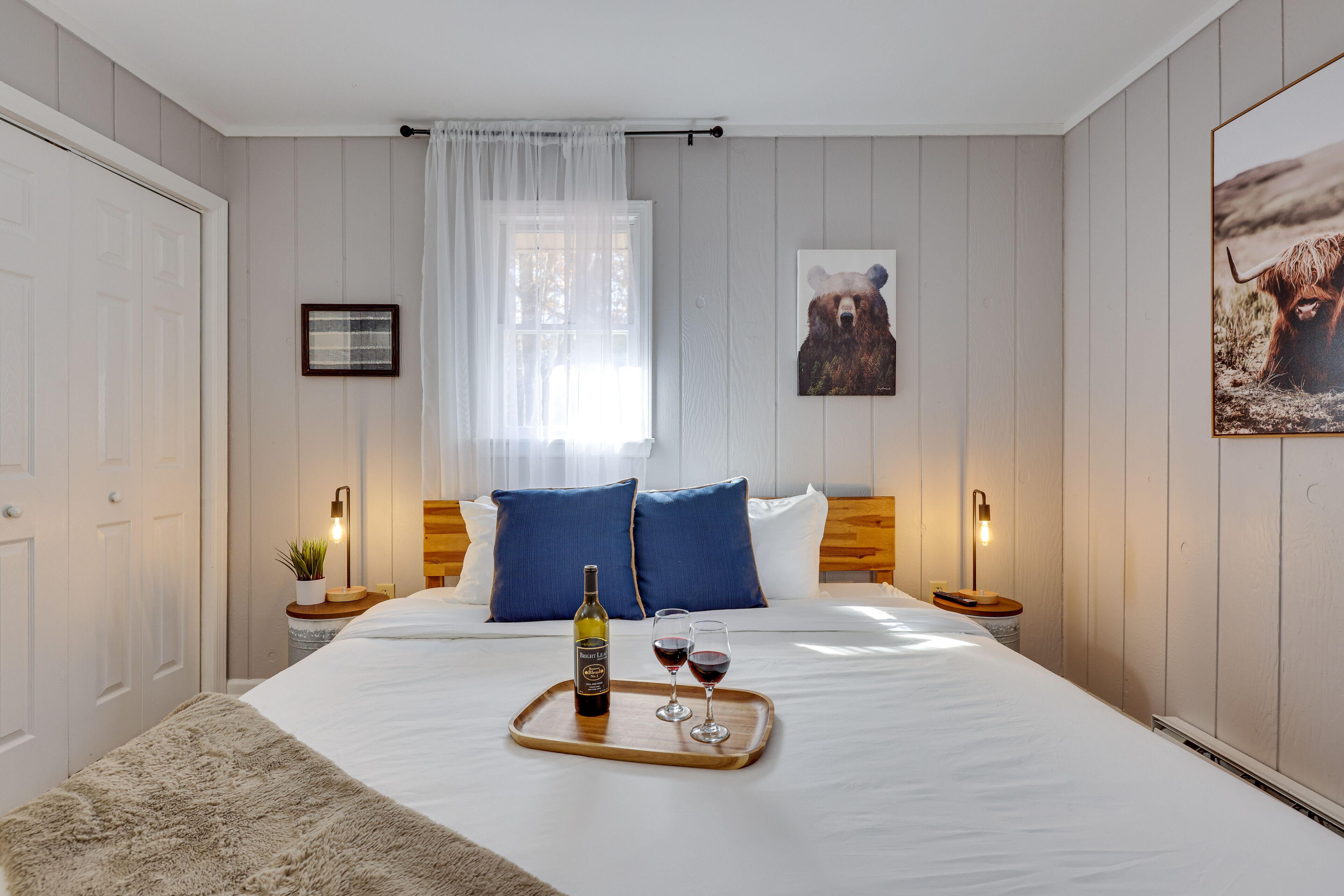 A bedroom with a large bed, two bedside tables, a bedside lamp, a window with sheer curtains, and a wall-mounted picture.