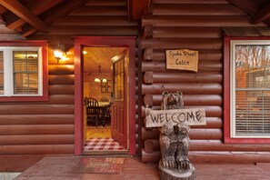 A wooden cabin with a 'Welcome' sign on a bear statue, a 'Spoke Wheel Cabin' sign, and a checkered mat at the entrance. - Long Mtn. Views - Covered Porch - Rockers - Fire Pit - Gated - Clean - 5 Stars! (Waynesville)