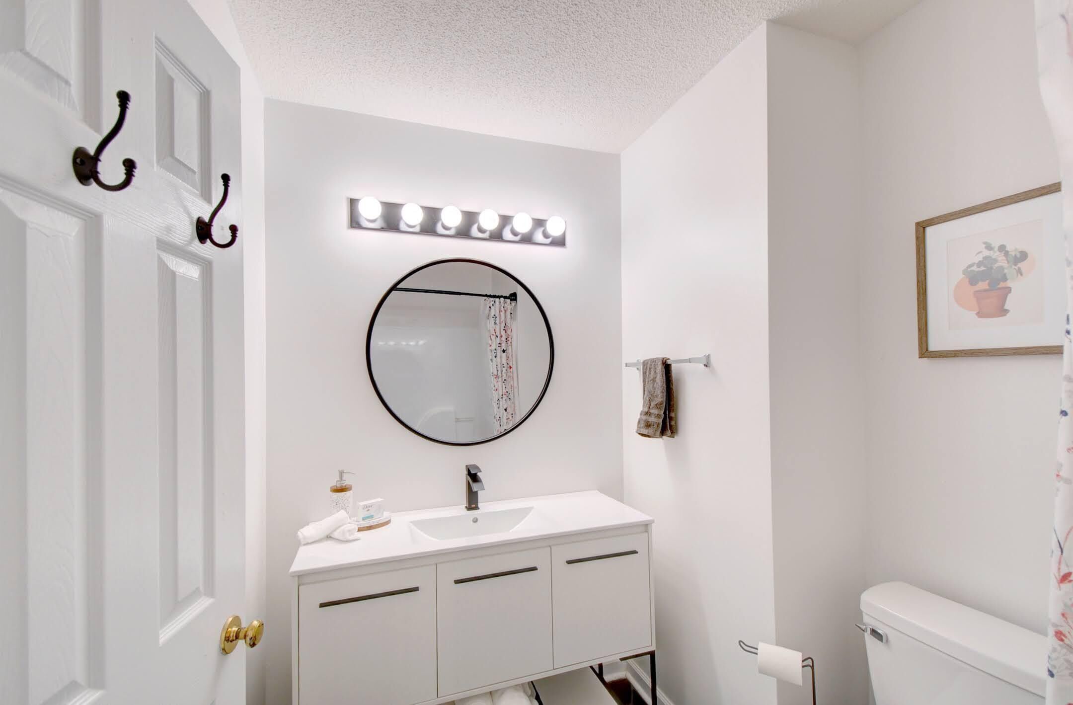 Un baño con tocador blanco, un espejo redondo con iluminación, un perchero y un cuadro en la pared.