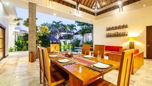 A dining area with a long wooden table set for four, surrounded by wooden chairs. A red sofa is visible in the background.