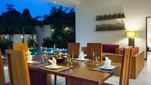 A dining area with a table set for six, surrounded by wooden chairs, with a view of a swimming pool and palm trees in the background.