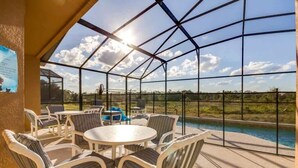 A covered outdoor patio with a pool view, featuring a round table and chairs, and a clear sky.