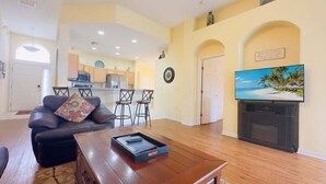 Un salón con sofá, mesa de centro y televisor. Se observa una cocina al fondo con zona de comedor.