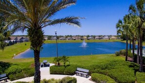 A serene lakeside view with a fountain, palm trees, and a row of houses in the background.