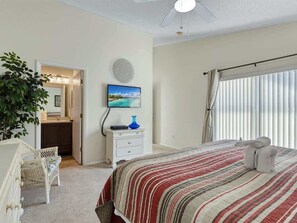 A bedroom with a bed, a TV, a dresser, a plant, and a window with blinds.
