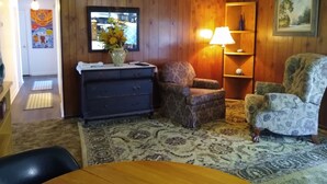 Ein Wohnzimmer mit Holzwand, einer Couch, einem gemusterten Sessel, einer Lampe, einer Vase mit Blumen und einem Gemälde.
