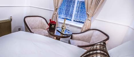 A room with a bed, two armchairs, a small table with a bottle and glass, and a window with curtains.