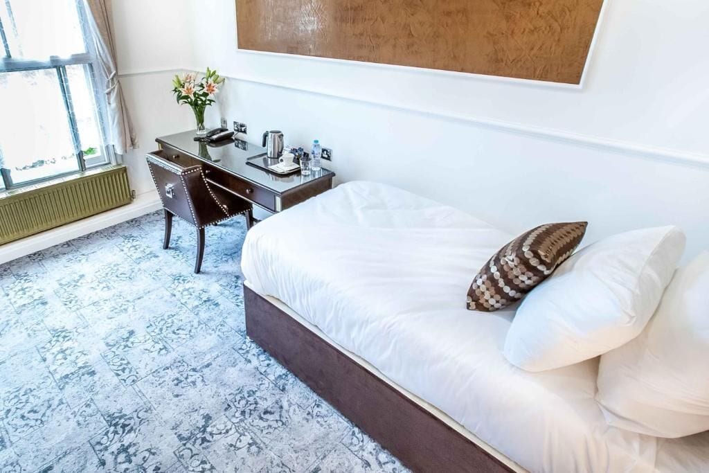 A hotel room with a bed, a desk, a chair, a radiator, and a window.