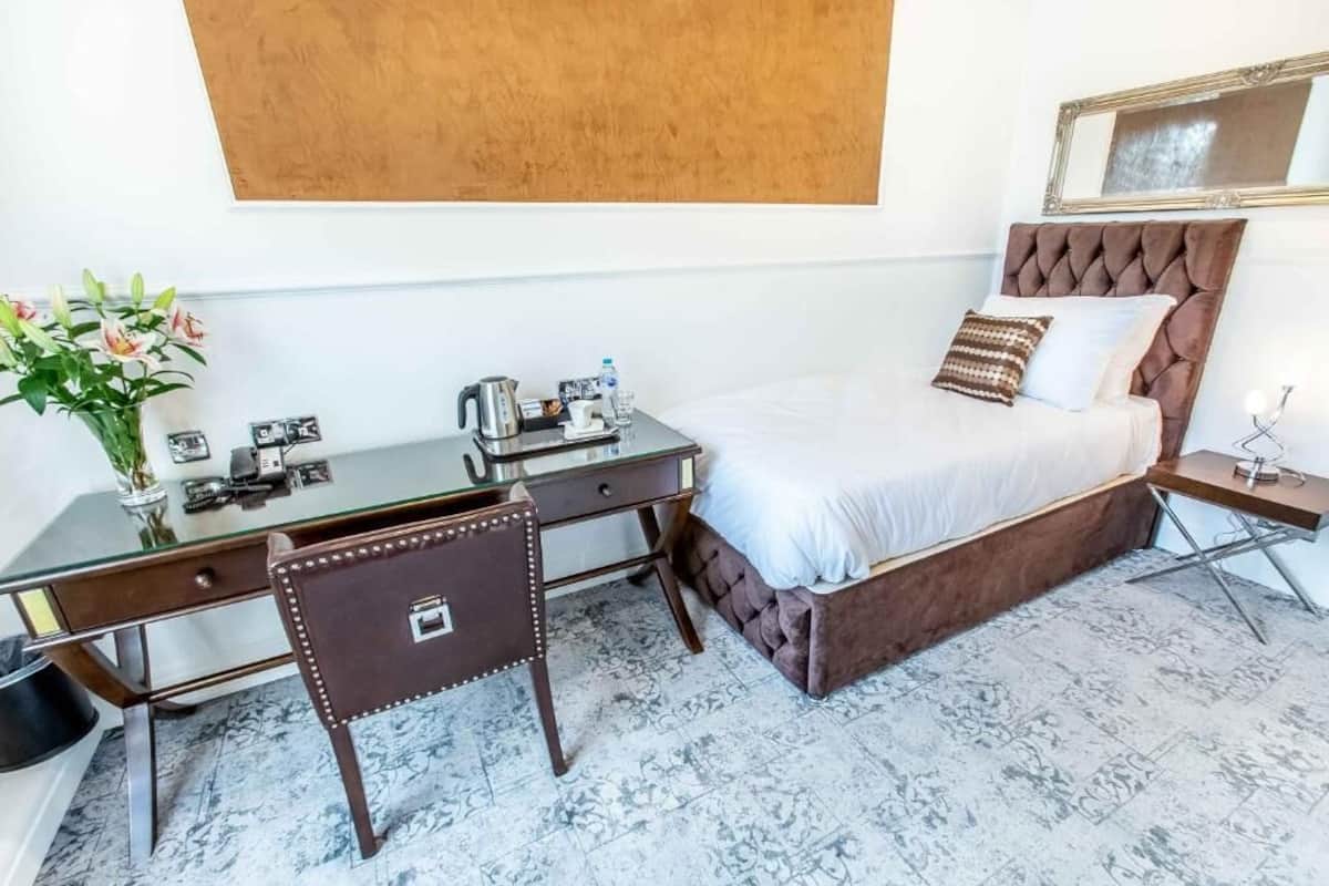 A hotel room with a bed, a desk, a chair, a small table, a mirror, and a vase with flowers.