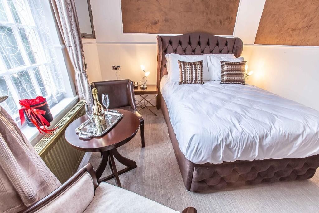 A hotel room with a large bed, a small table with a bottle and glasses, a chair, and a window with curtains.