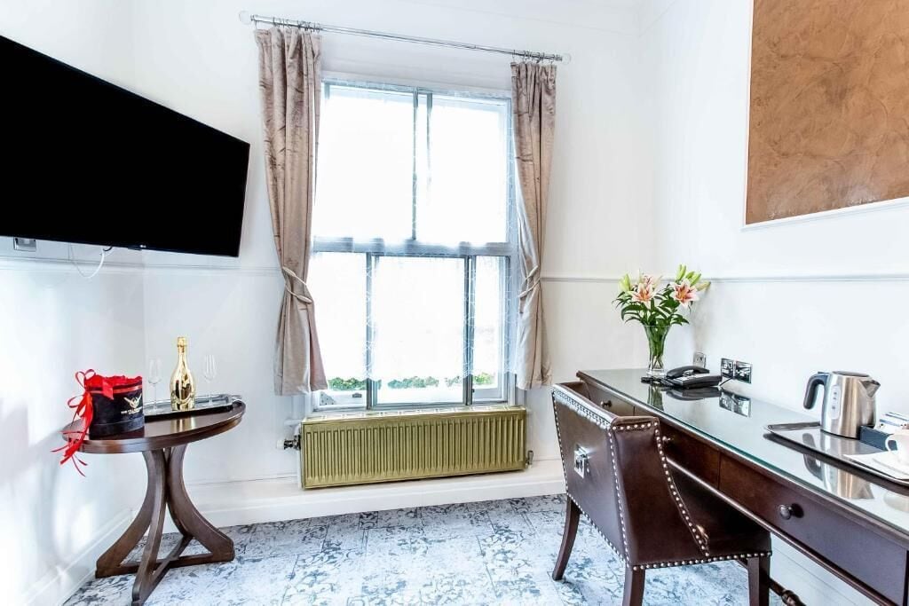 Quarto com uma janela ampla, radiador, escrivaninha com cadeira, televisão de tela plana e um vaso com flores.