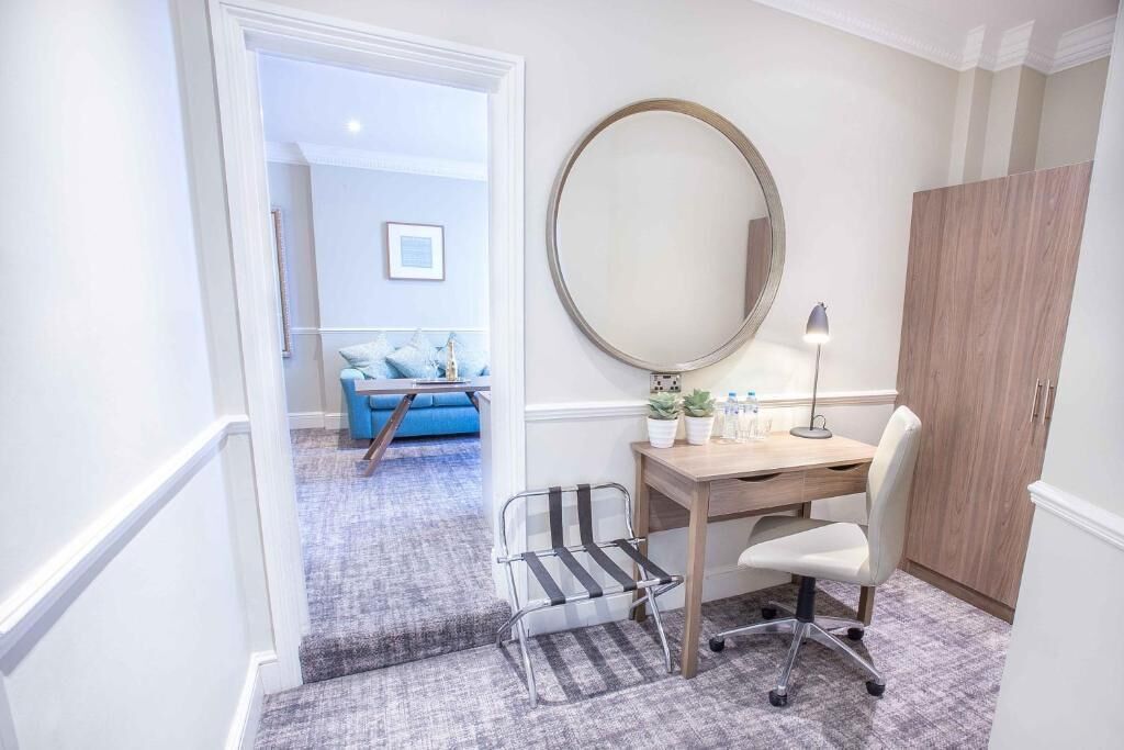 A room with a desk, chair, and a large mirror. There is a blue sofa and a small table with plants in the background.