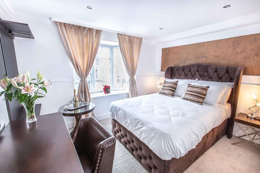 Habitación de hotel con una cama grande, un escritorio, una silla, una mesita con un jarrón de flores y una ventana con cortinas.