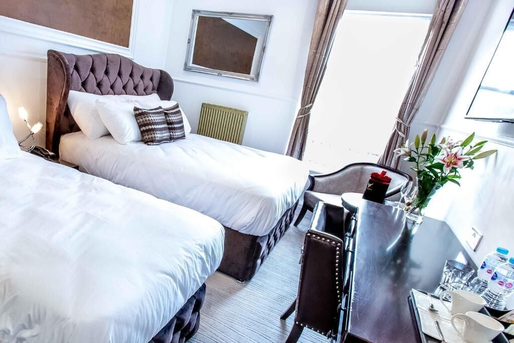 A hotel room with two beds, a desk with a chair, a mirror, a radiator, and a window with curtains.