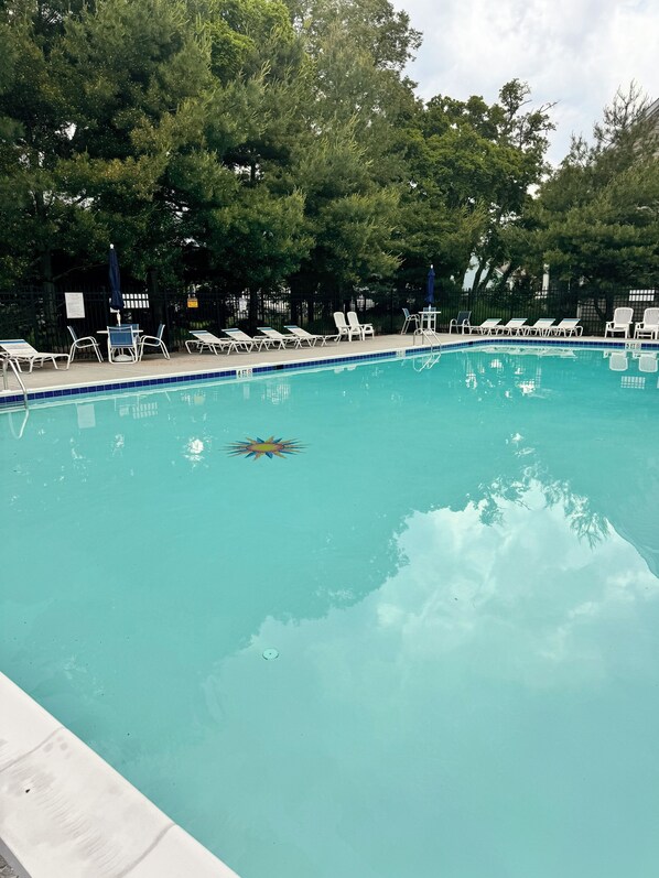 A swimming pool with clear blue water, surrounded by lounge chairs and umbrellas, and trees in the background.