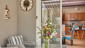 Une table à plateau en verre sur laquelle est posé un vase de fleurs, des chaises en osier, une horloge, et une vue sur un coin cuisine.