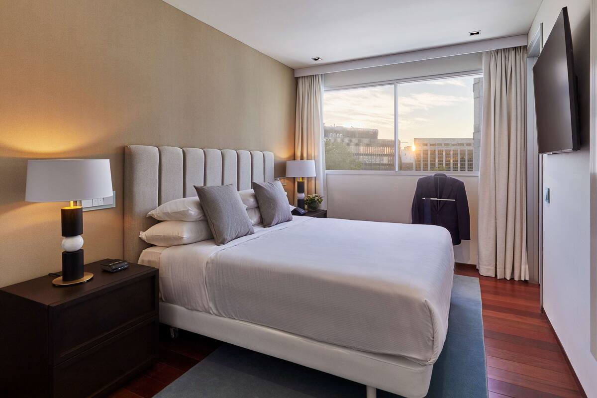 Una habitación de hotel moderna con una cama grande, dos mesitas de noche, un televisor de pantalla plana y vistas a la ciudad desde la ventana.