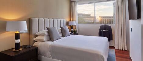 A modern hotel room with a large bed, two bedside tables, a flat-screen TV, and a view of the cityscape through the window.