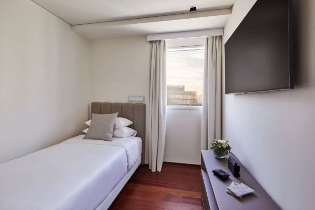 A hotel room with a single bed, a small table with a remote control, and a TV mounted on the wall.