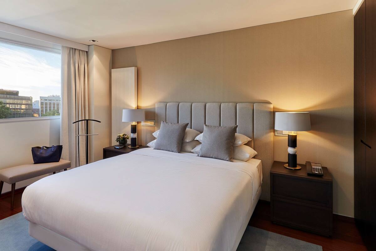Habitación de hotel moderna con una cama grande, dos mesitas de noche con lámparas, un banco y vistas a la ciudad.