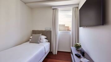 Habitació d'hotel amb un llit individual, un escriptori petit amb una làmpada, una televisió de pantalla plana i una finestra amb cortines.